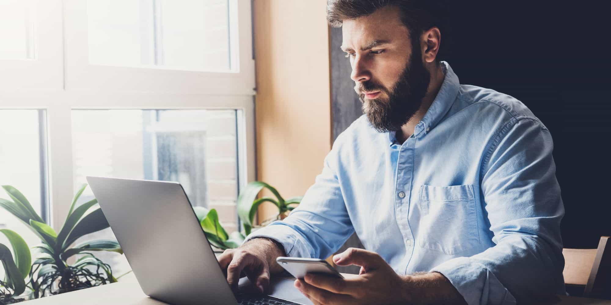 Mężczyzna siedzący przy laptopie ze smartfonem w ręce zlecający przelewy walutowe.